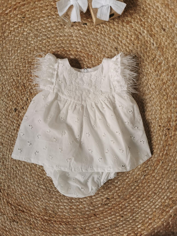A white baby dress with lace trim and feather details is displayed on a woven mat.