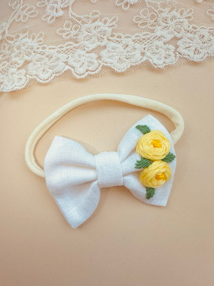 A white bow with yellow flowers is displayed on a beige background, accompanied by a lace doily.