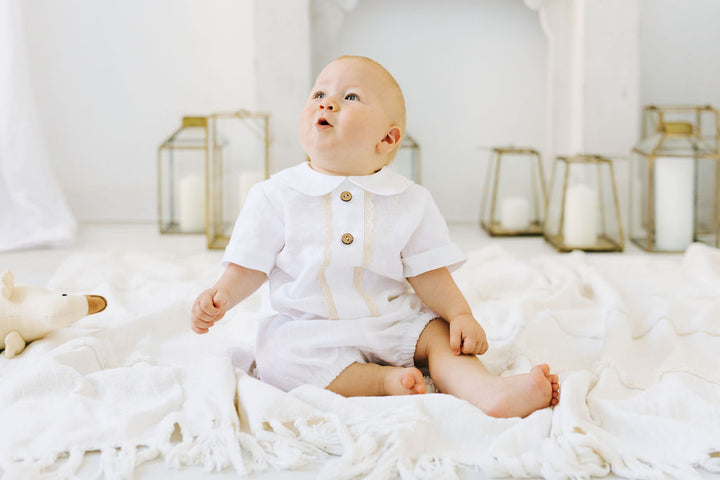 A baby in a white outfit sits on a white blanket, looking up with curiosity.