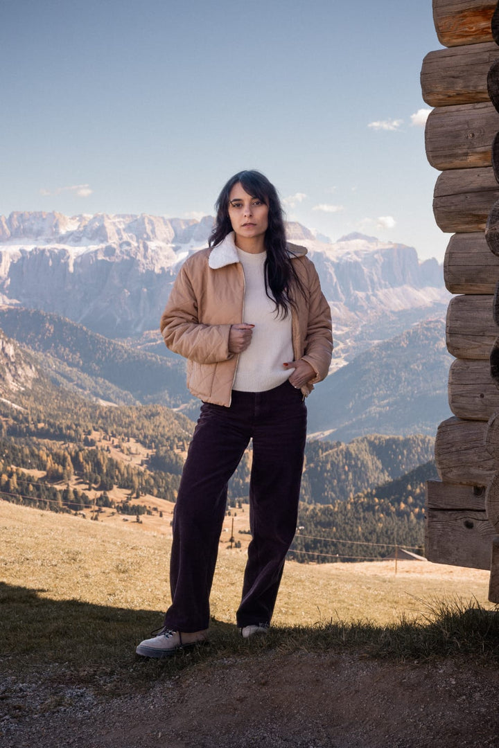 A woman in a beige jacket stands in front of a wooden cabin, with a mountainous landscape in the background.