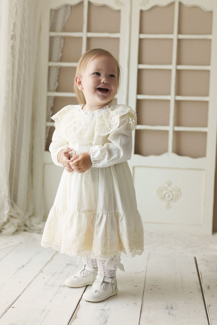 A young girl in a white lace dress stands in front of a white door, smiling and looking up.