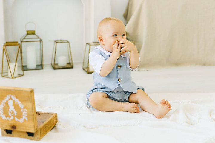 A baby sitting on a white blanket, wearing a blue outfit and holding a toy in their mouth.