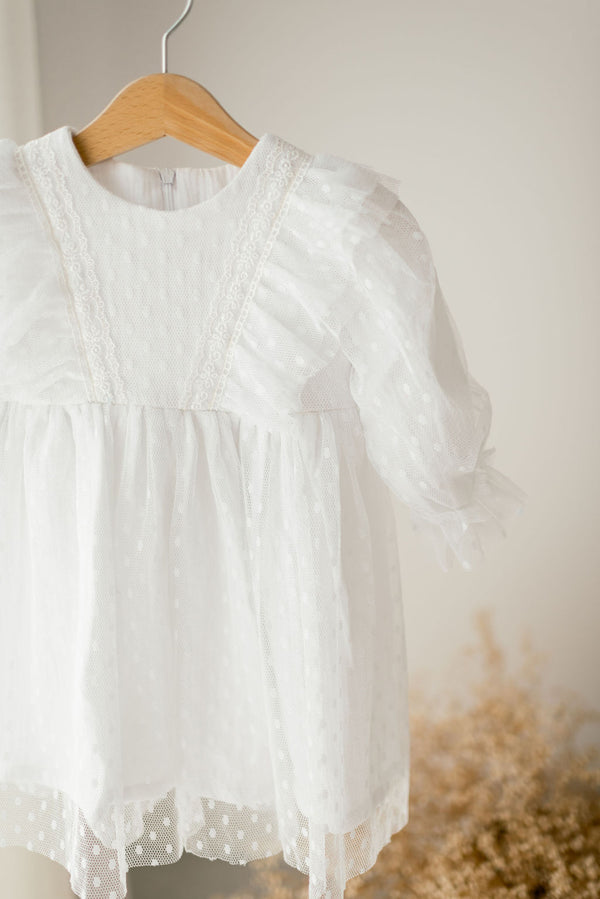 A white, ruffled dress with lace trim hangs on a wooden hanger against a light background.