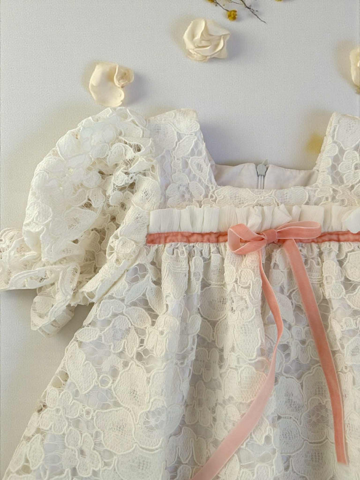 A white lace dress with a pink ribbon, placed on a white surface with small white flowers scattered around it.