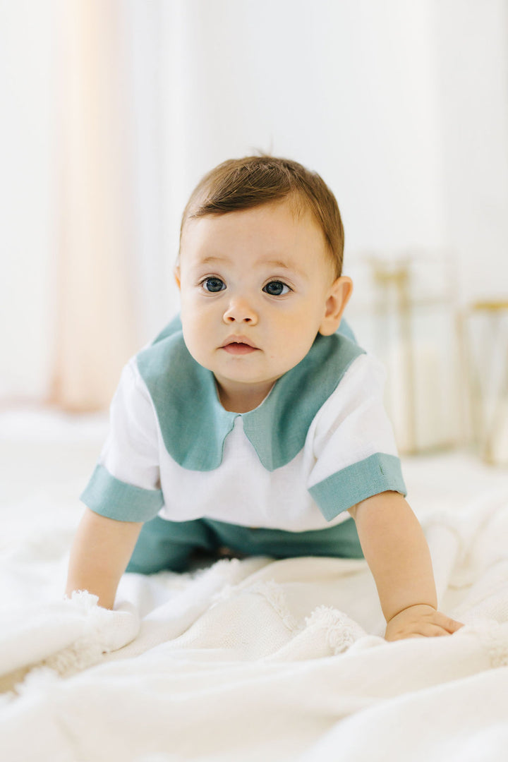 A baby boy in a white and green outfit is sitting on a white blanket, looking directly at the camera with a thoughtful expression.