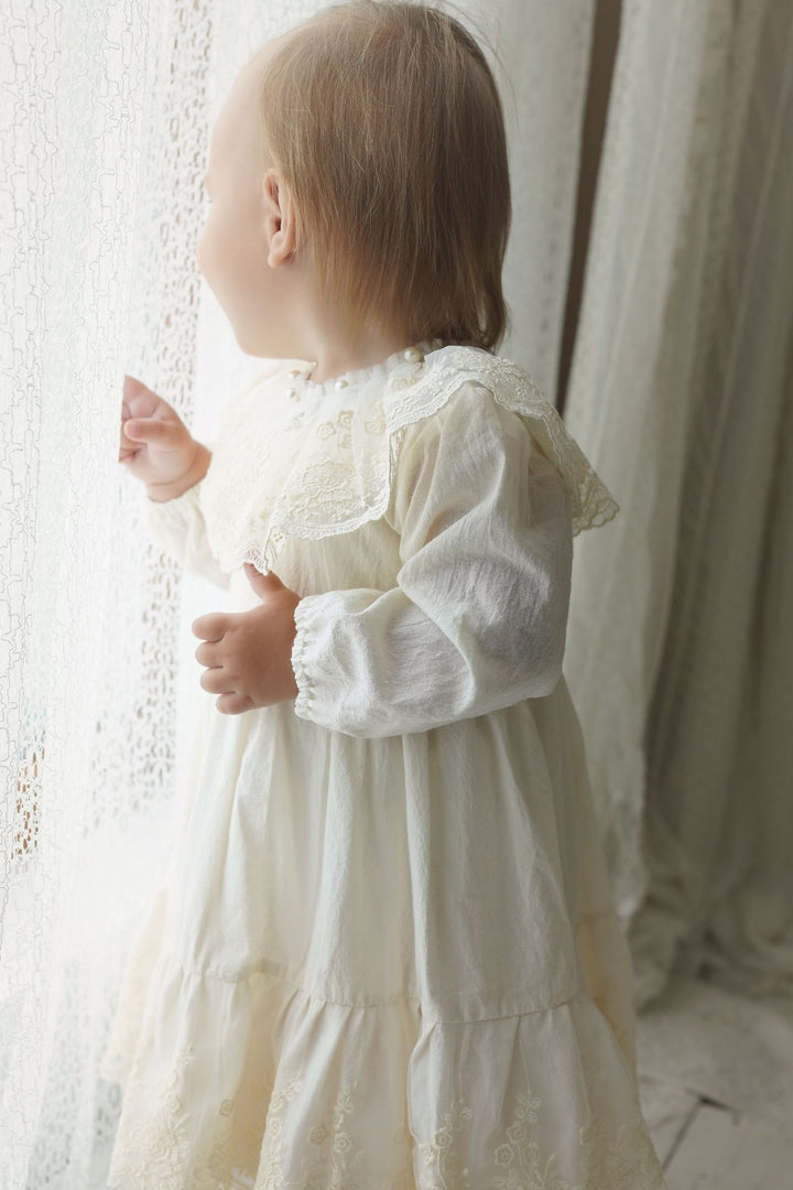 A young child in a white lace dress stands in front of a window, gazing out with curiosity.
