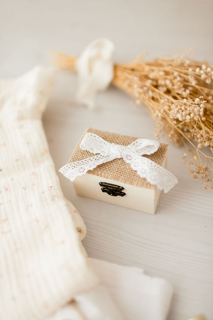 A small wooden box with a white lace bow tied around it, placed on a white surface next to dried flowers and a white fabric.