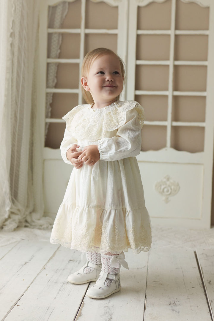 A young girl in a white lace dress stands on a wooden floor, smiling and looking up.