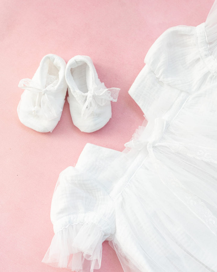 A pair of white baby shoes and a white dress with lace trim are placed on a pink background.