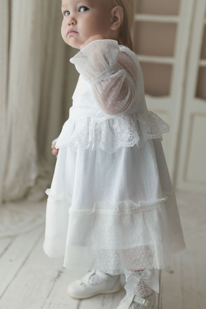 A young child in a white lace dress stands in a room with white curtains.