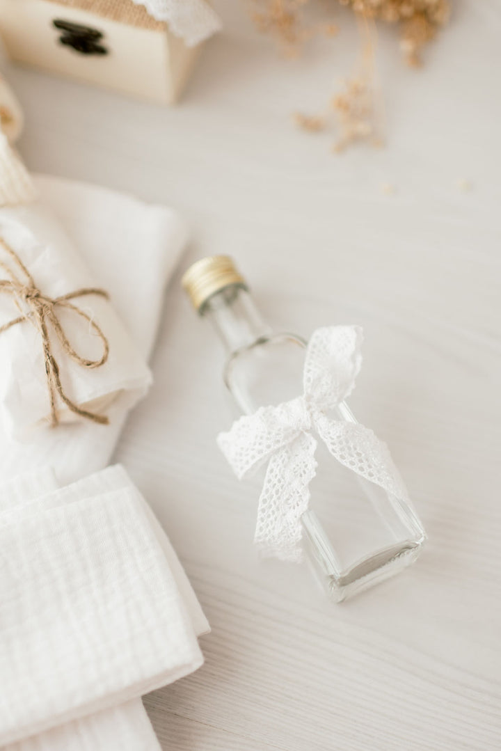 A small glass bottle with a white lace bow tied around it, placed on a white surface alongside other items such as a small wooden box and a white cloth.