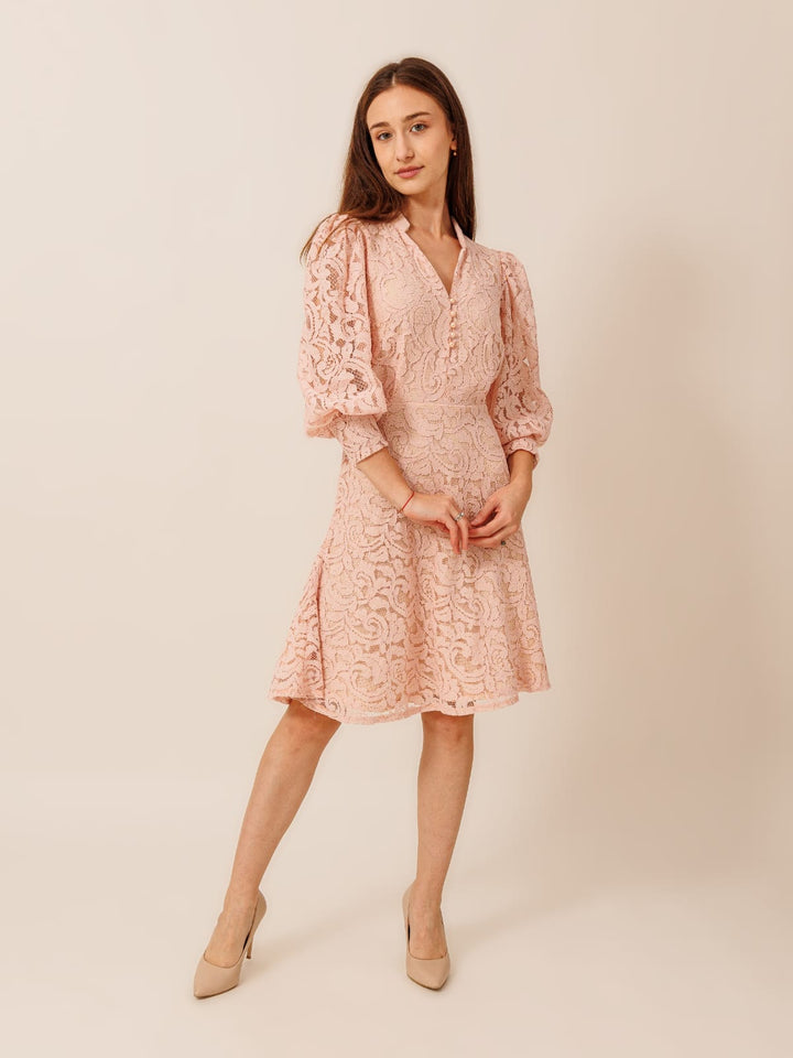 A young woman wearing a pink lace dress with puffed sleeves and a flared skirt, posing confidently against a plain background.