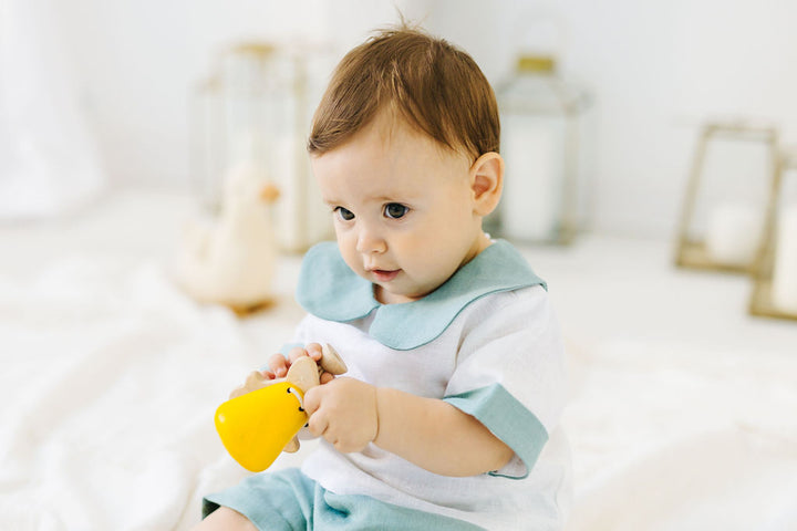 A baby boy in a white shirt and blue shorts is sitting on a white bed, holding a yellow toy and looking at the camera.