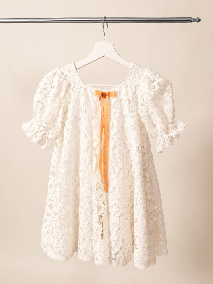 A white lace dress with a single orange ribbon tied in a bow, hanging on a clothing rack.