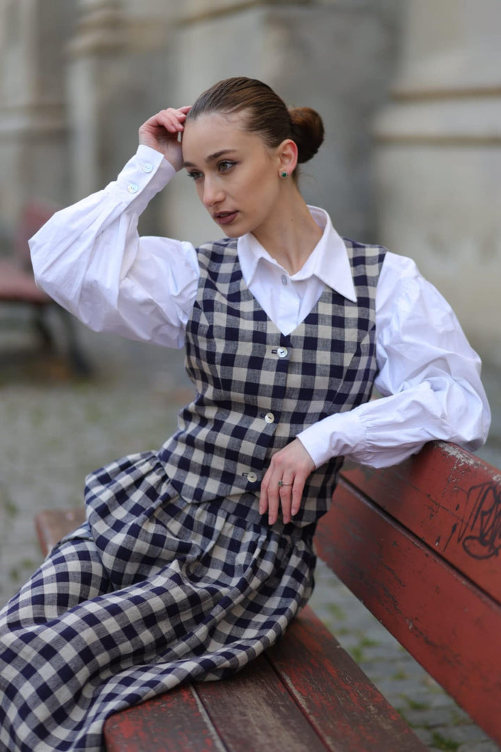 A young woman in a white shirt and blue and white checkered dress sits on a wooden bench, her hand resting on her head.