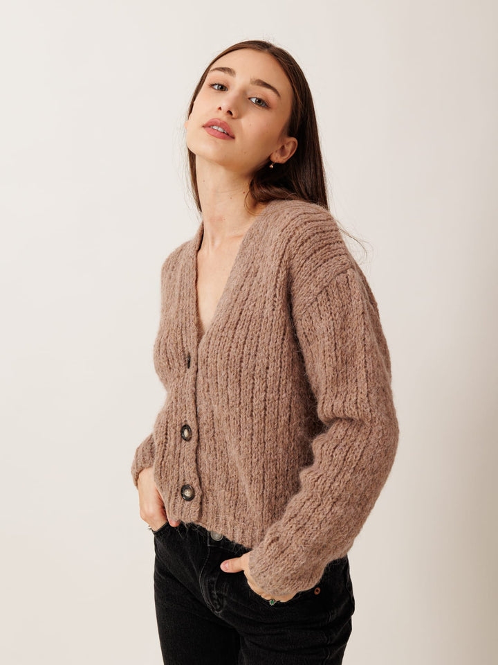 A woman wearing a brown cardigan with a v-neck and button-down front, posing with her hands in her pockets against a plain white background.