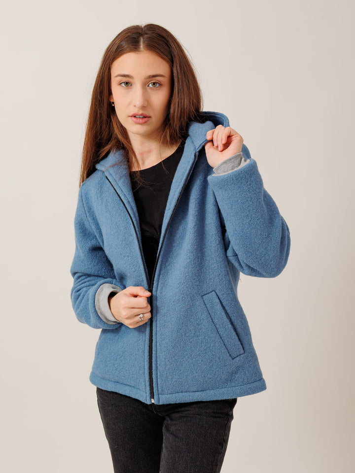 A young woman with long brown hair wearing a blue hooded jacket stands against a plain background, looking directly at the camera with a serious expression.