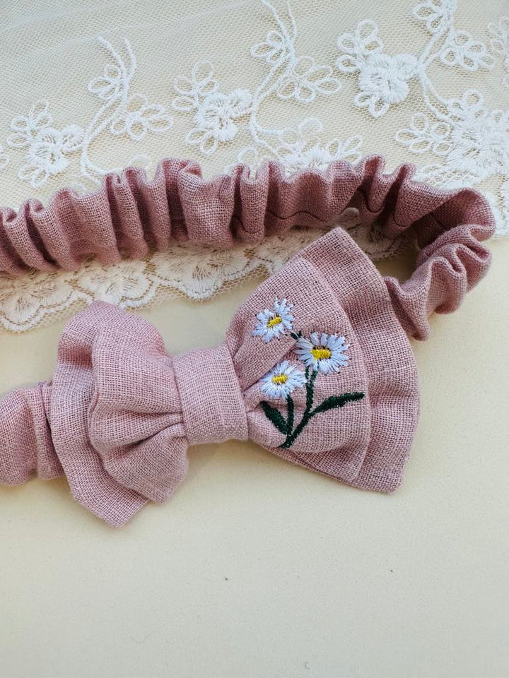 A pink bow tie with white daisies embroidered on it, resting on a white lace background.