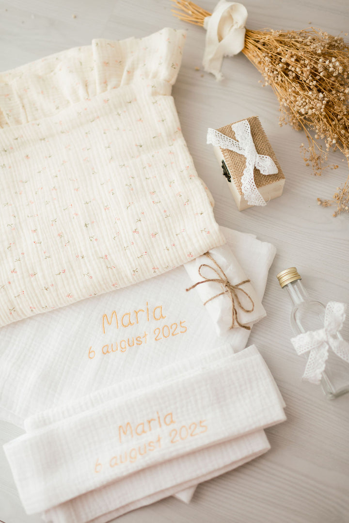 a collection of items, including a white towel with the name "Maria" printed on it, a small box with a ribbon, a bottle, and some dried flowers, all arranged on a white surface.