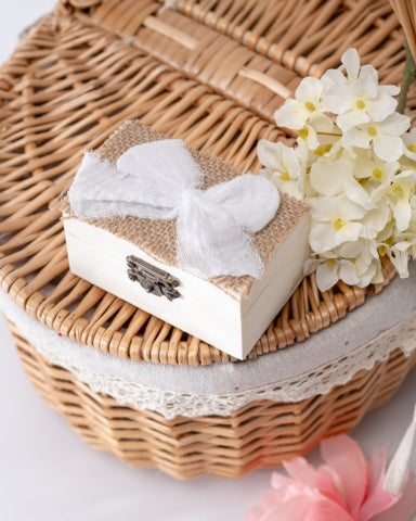 A small white wooden box with a bow on top, placed on a wicker basket with a white lace trim.