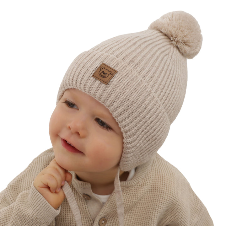 A young child wearing a beige knit hat with a pom pom on top, smiling at the camera.