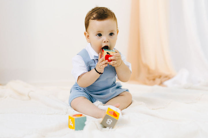 A baby boy sitting on a white blanket, holding a toy in his mouth.