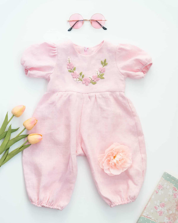 A pink floral jumpsuit with a flower on the front, pink pom poms, and pink sunglasses are arranged on a white background.