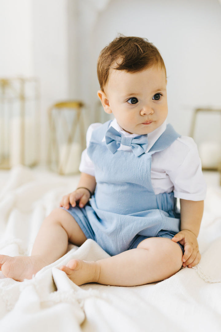 A baby boy wearing a blue dress and bow tie sits on a white blanket, looking directly at the camera with a thoughtful expression.