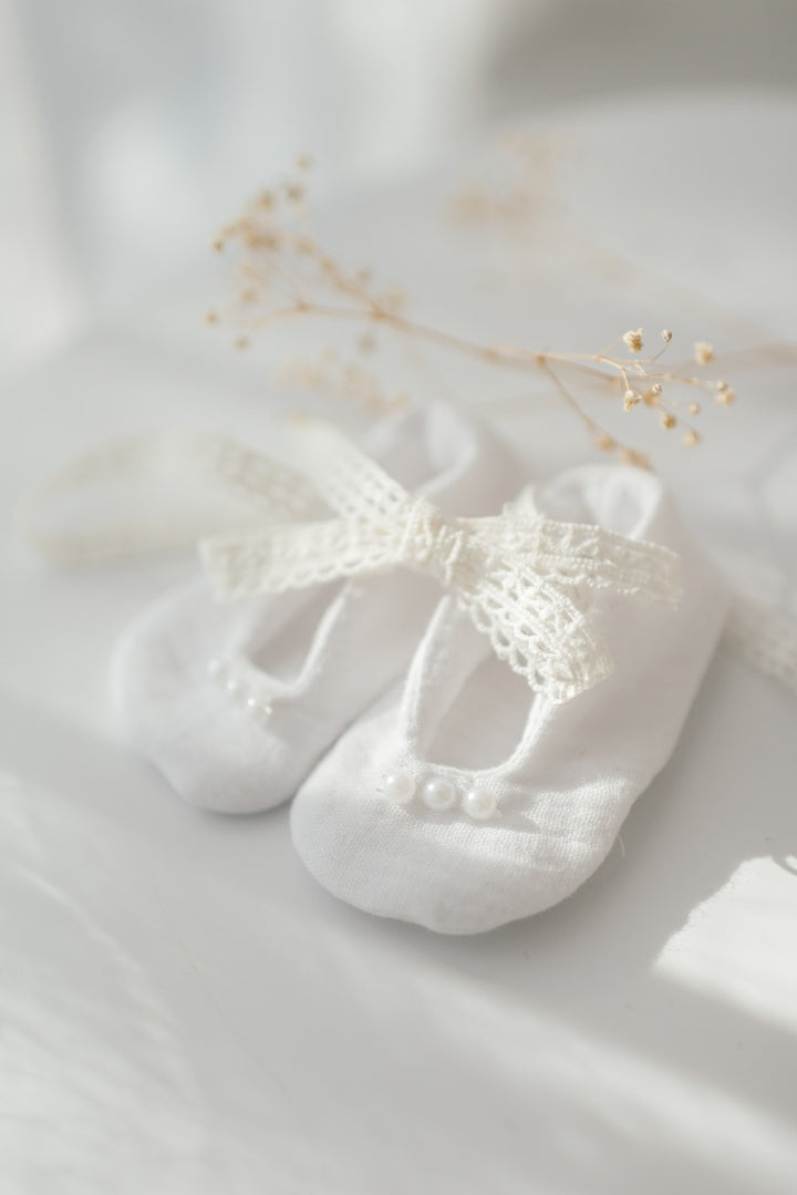 A pair of white baby shoes with a delicate lace bow sits on a white surface, with delicate white flowers or baby's breath sprouting from the top.