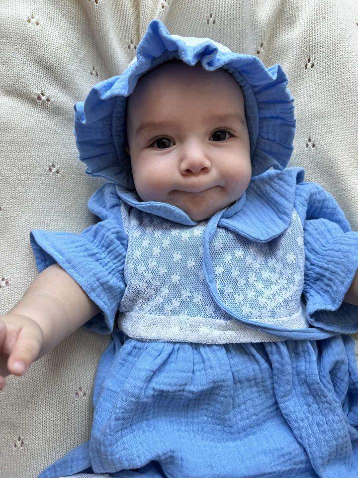 A smiling baby wearing a blue dress and a blue bonnet lies on a white blanket.