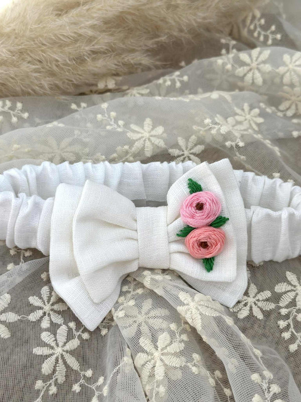 A white headband with a bow and two pink flowers is displayed on a white lace background.
