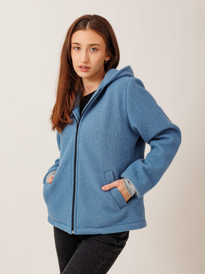 A young woman with long brown hair wearing a blue hooded jacket stands against a plain background.