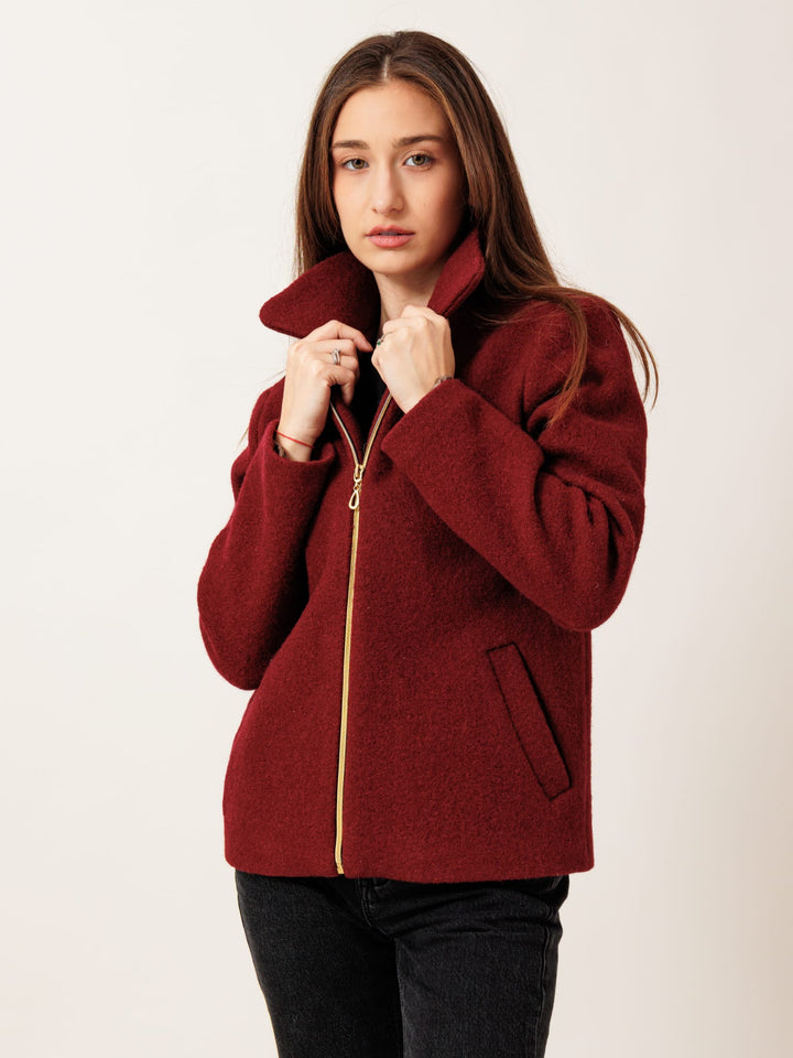 A young woman wearing a red jacket with a gold zipper stands against a plain background, looking directly at the camera with a serious expression.