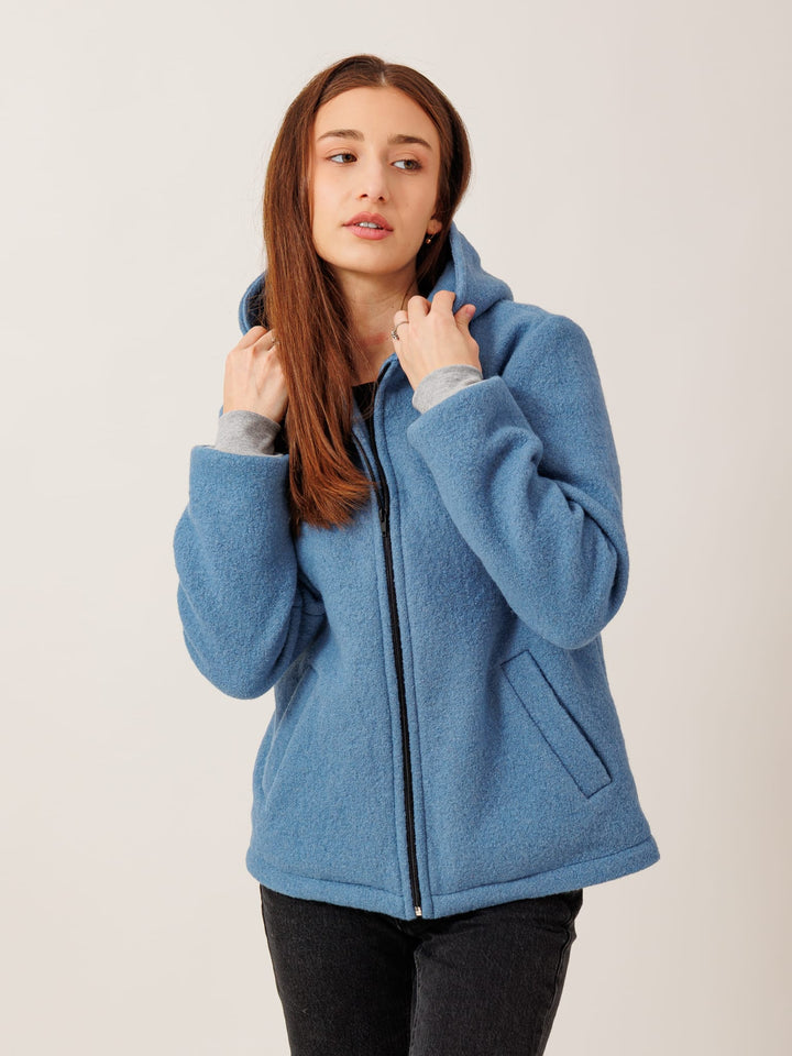 A young woman with long brown hair wearing a blue hooded jacket stands against a plain background.