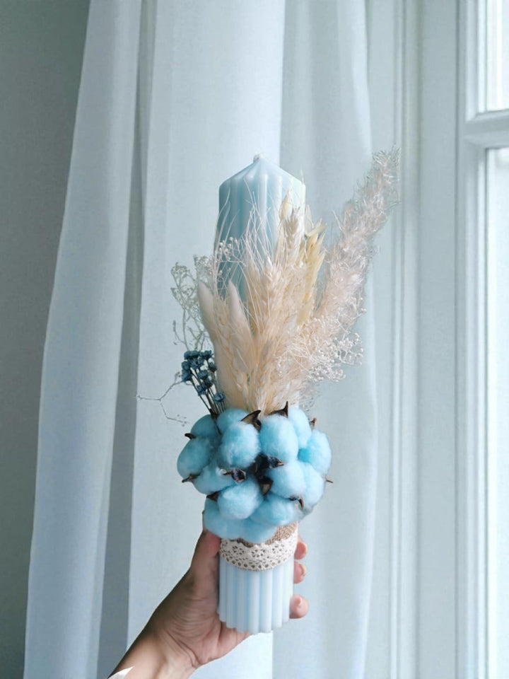 A hand holding a white vase filled with blue cotton balls and dried flowers, placed in front of a white curtain.