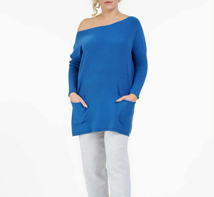 A woman wearing a blue off-the-shoulder sweater and gray pants stands against a white background.