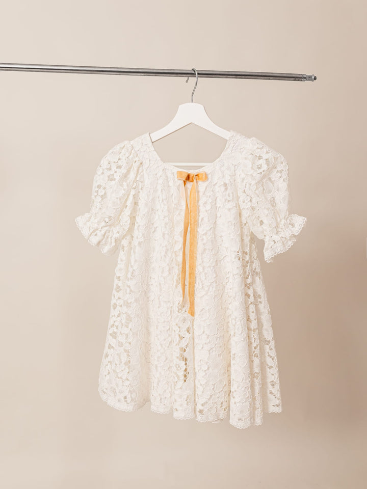 A white lace dress with a yellow ribbon is hanging on a clothing rack.