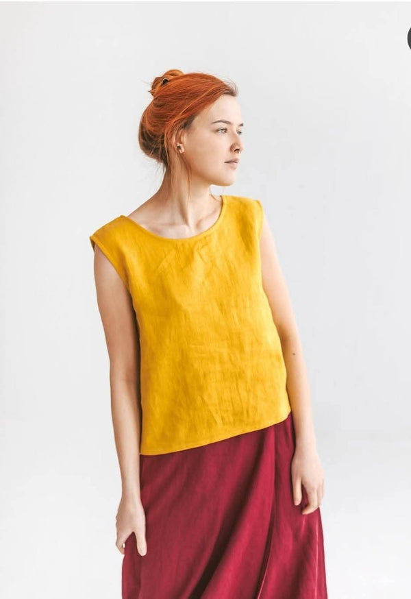 A woman with red hair wearing a yellow sleeveless top and a red skirt stands against a white background.