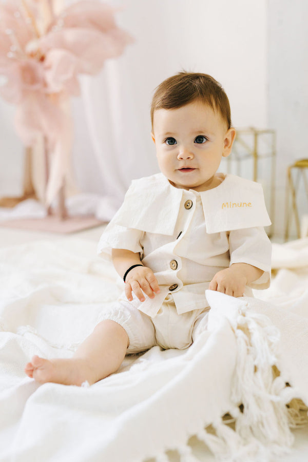 A baby dressed in a white outfit sits on a white blanket, with a pink flower in the background.