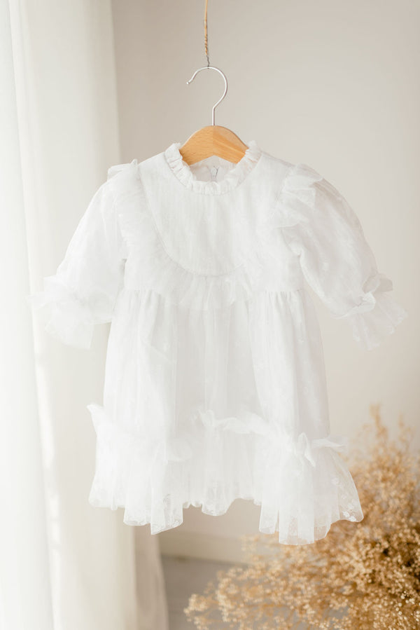 A white, ruffled dress is hanging on a wooden hanger against a white wall.