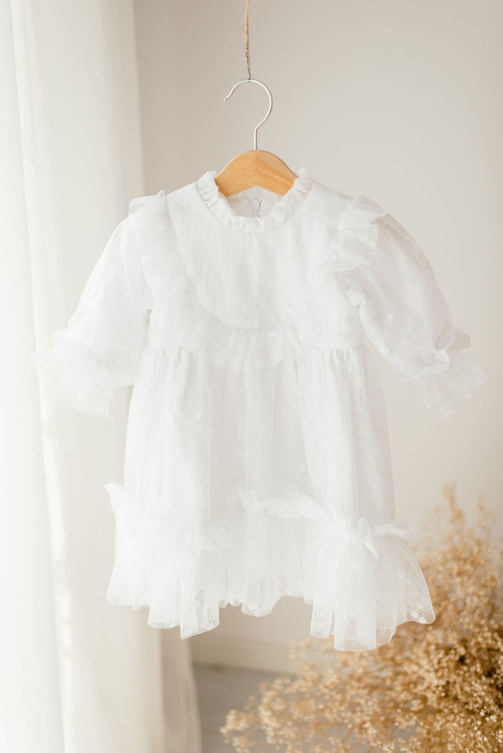 A white, ruffled dress is hanging on a wooden hanger against a white wall.