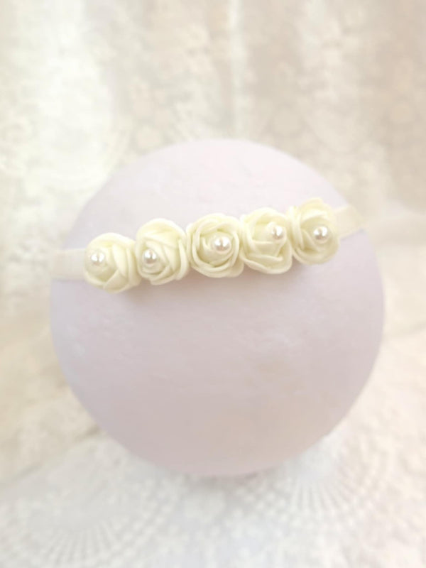 A white headband adorned with white roses and pearls is displayed against a white background.