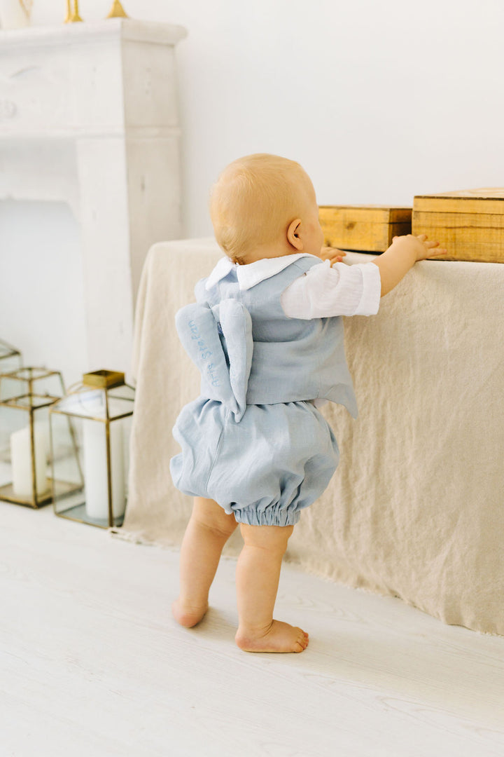A baby in a light blue outfit is standing on a white floor, reaching up to touch a wooden box on a shelf.