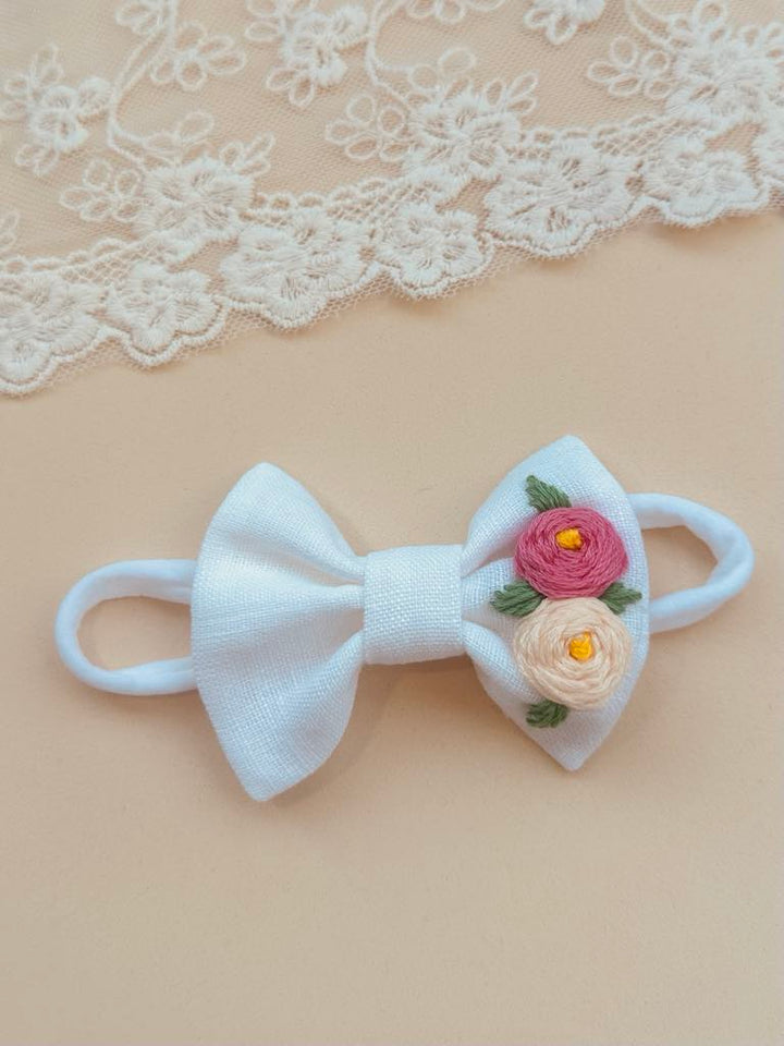 A white bow with pink and yellow flowers is placed on a beige background, with a lace doily visible in the top left corner.