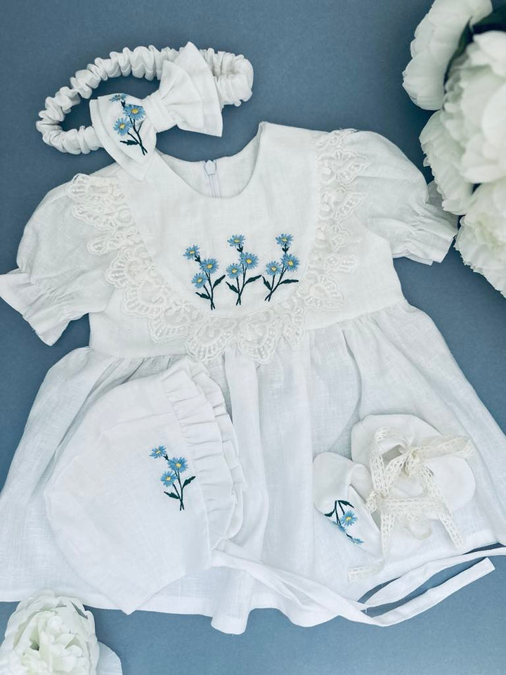 A white dress with blue flowers embroidered on it, along with a matching headband and a pair of white baby booties, all arranged on a blue background.