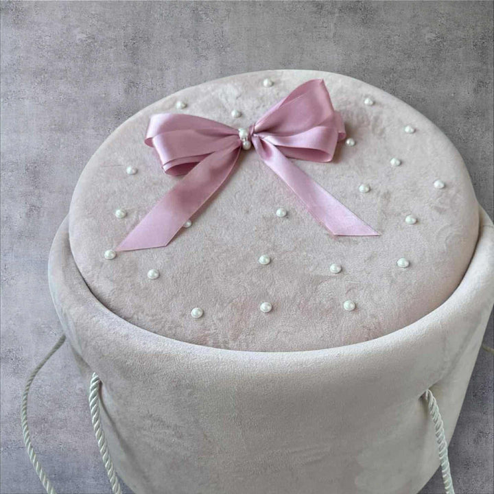 A round, white velvet box with a pink ribbon and white pearls on top, placed on a gray background.