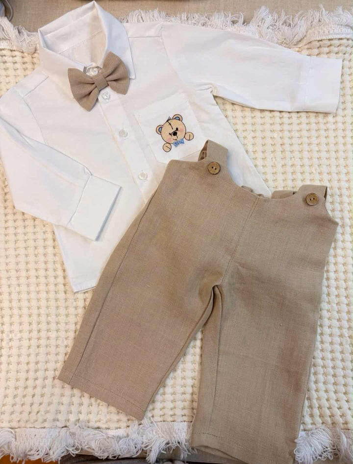 A white shirt with a bow tie and beige pants are laid out on a white surface, possibly a blanket or bed.