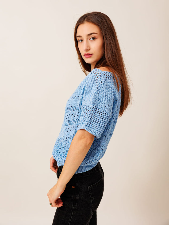 A young woman with long brown hair wearing a blue knitted sweater and black pants stands against a plain background, looking directly at the camera with a thoughtful expression.