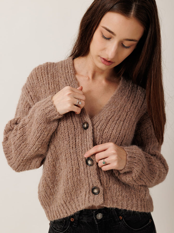 A woman wearing a brown cardigan stands against a white background, adjusting the buttons of her sweater.