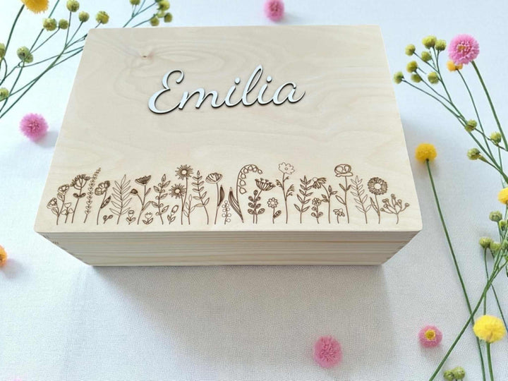 A wooden box with the name "Emilia" engraved on it, surrounded by various flowers and plants.